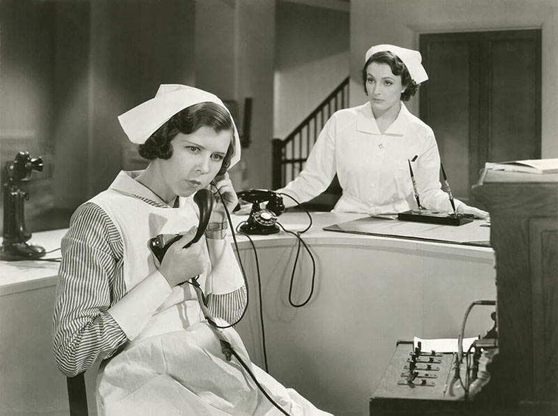 Two nurses, black and white image, one nurse on the phone behind a desk and the other nurse watching her in front of the desk