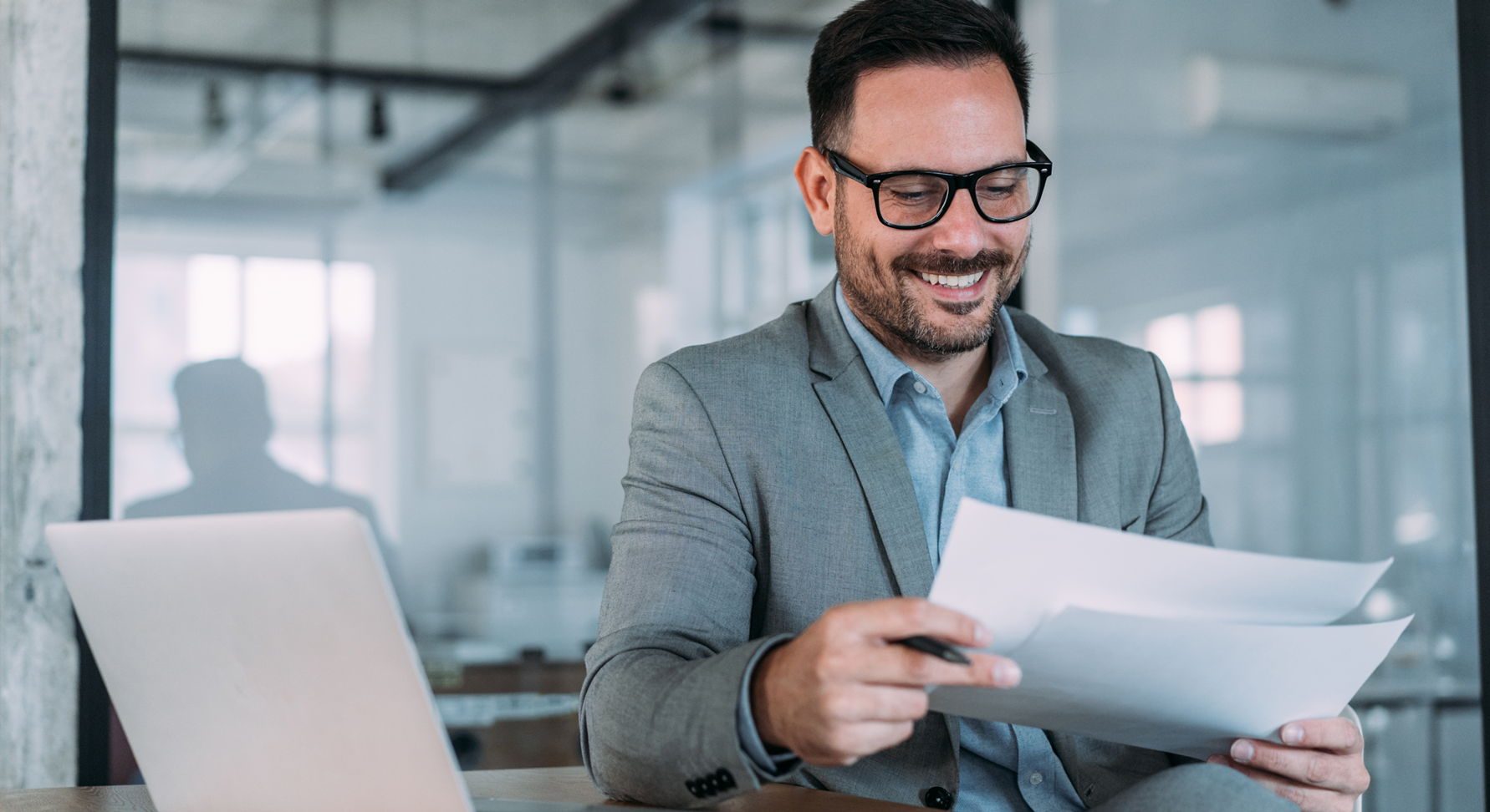 A smiling person holding a piece of paper in front of a laptop