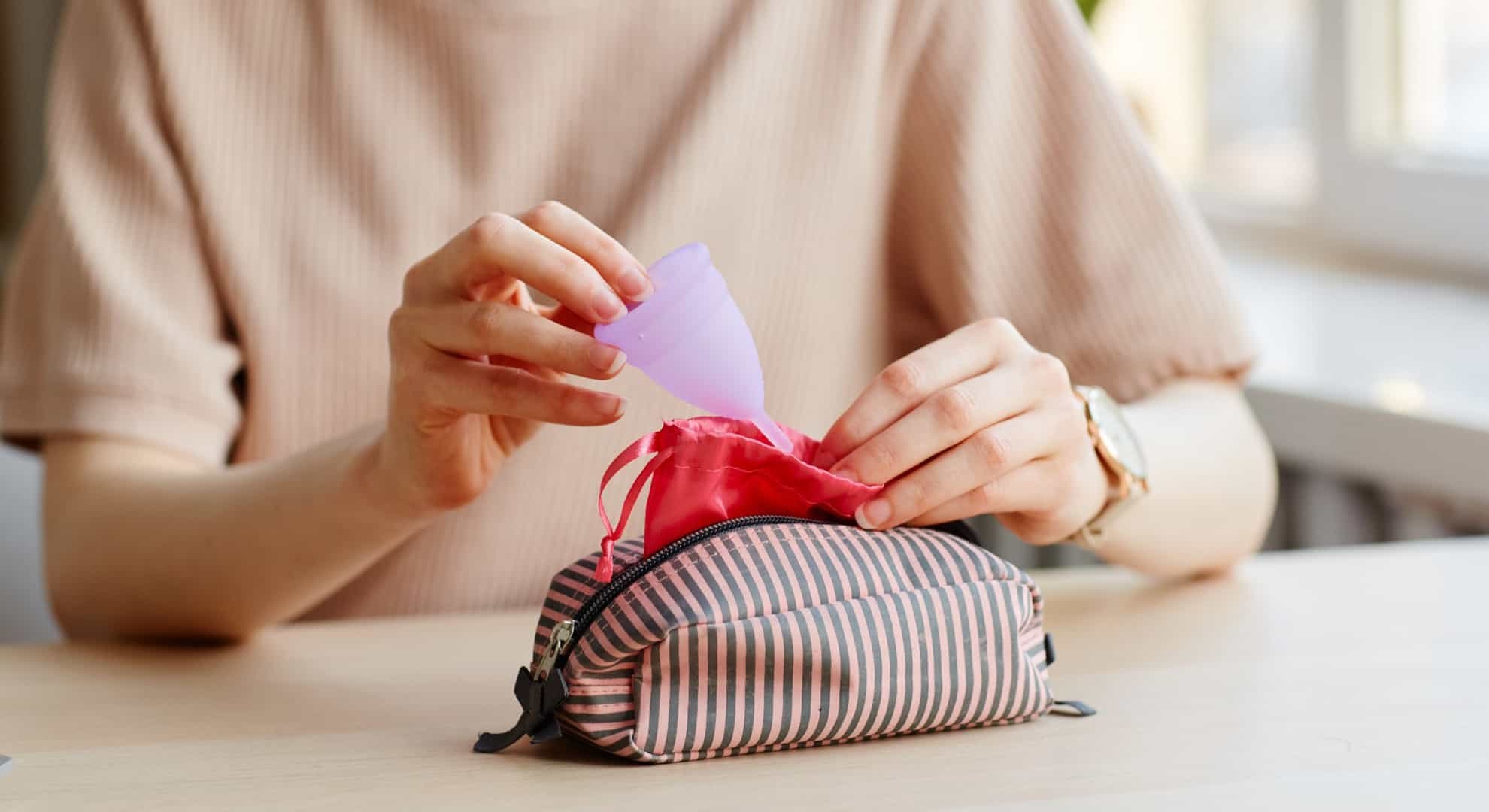 Closeup of young woman putting menstrual cup in cosmetic bag with other feminine essentials, copy space