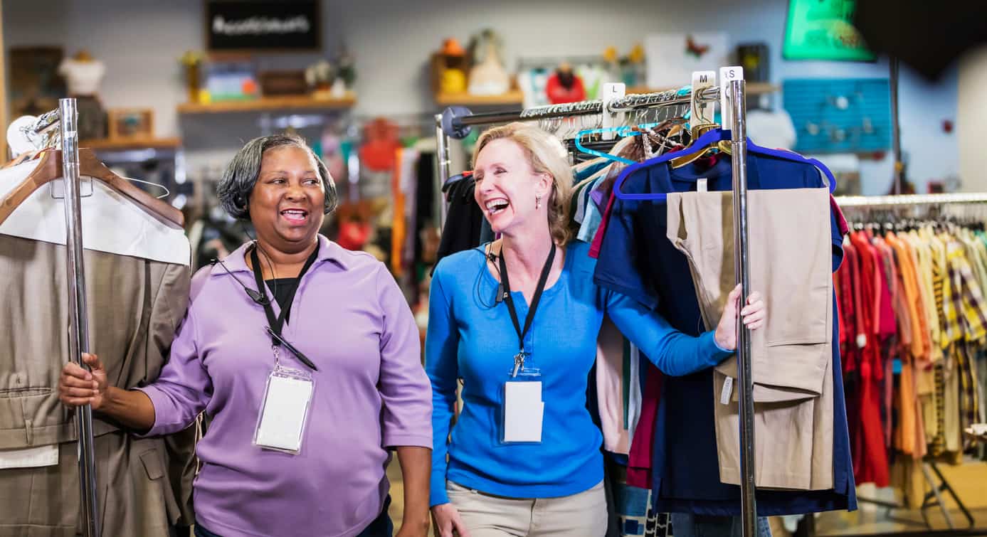 two women pulling racks of clothing through a store, talking to each other as they walk