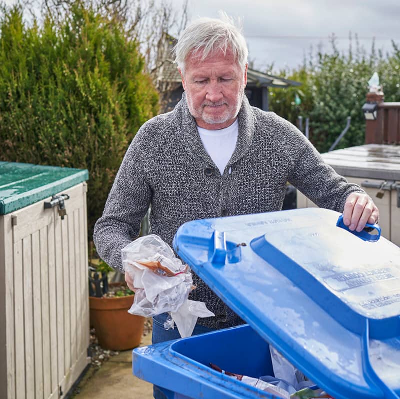 Photo of a man placing soft plastics into a blue recycling bin outside in a garden