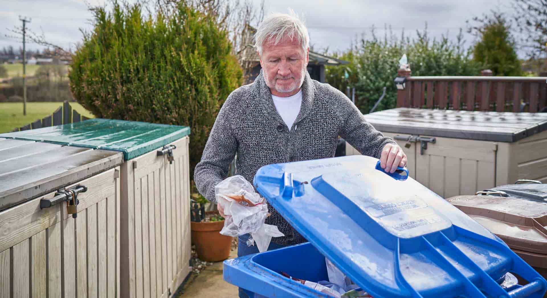 Photo of a man placing soft plastics into a blue recycling bin outside in a garden