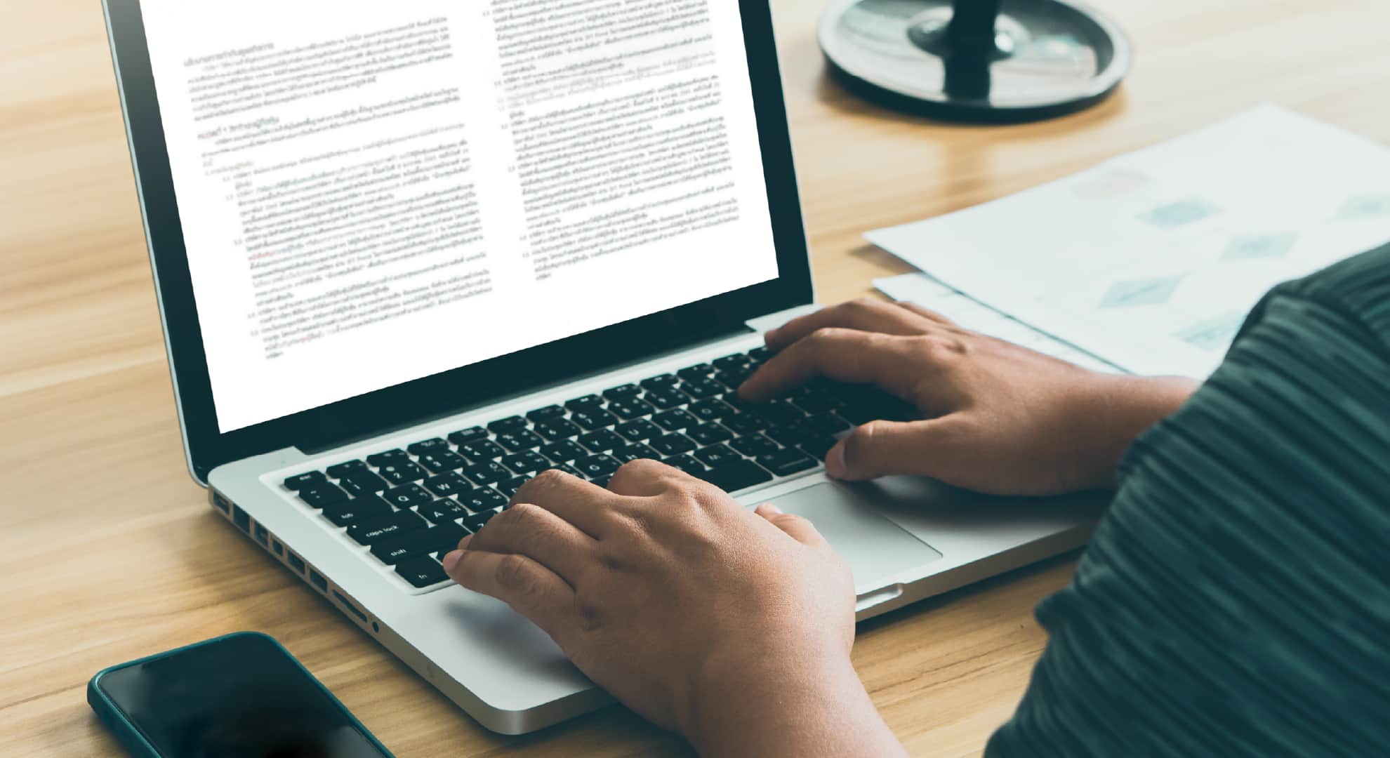 Person typing on a laptop at a wooden desk, with papers and a smartphone nearby.