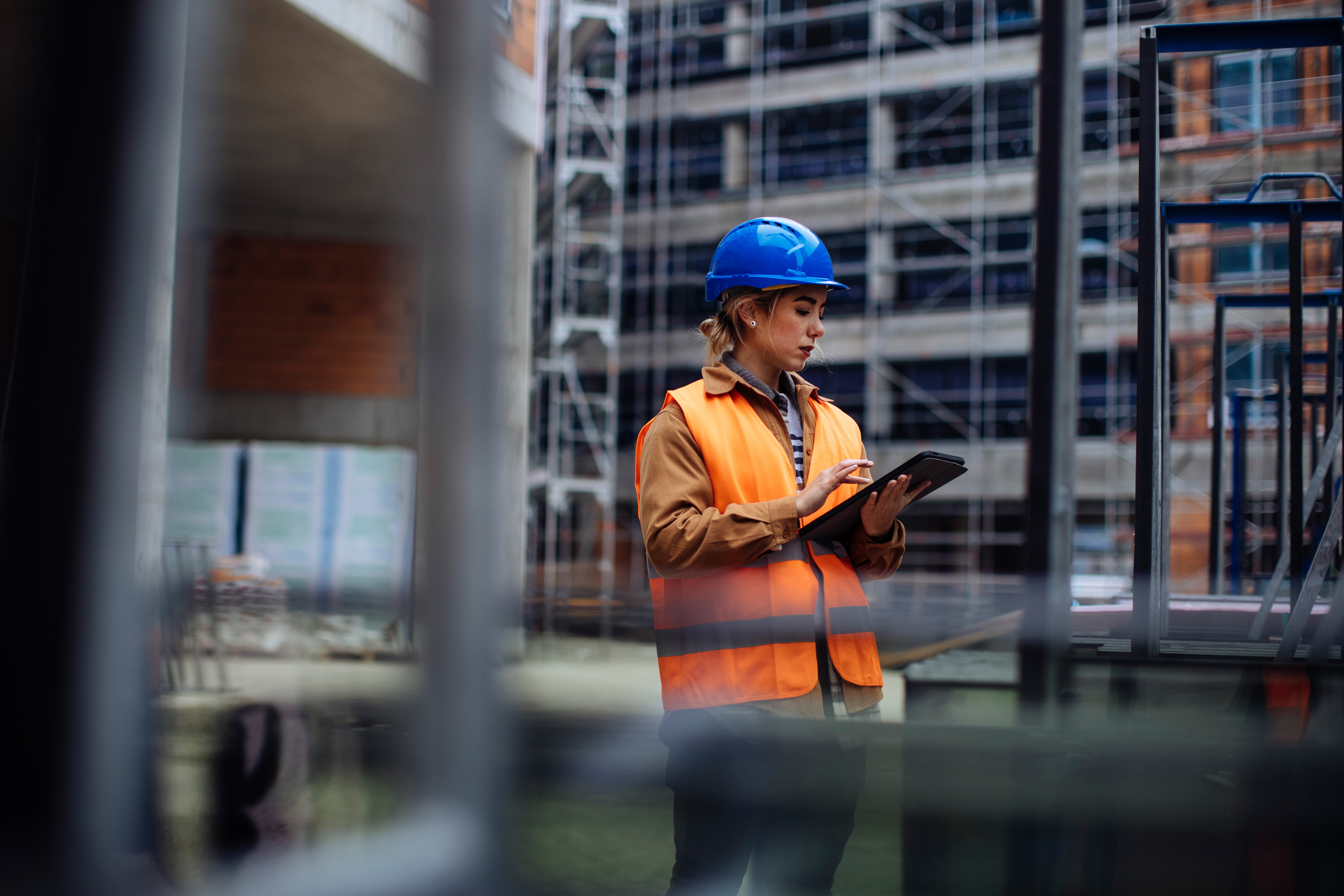 Image of a construction worker in blue hard hat and orange hi-vis with clipboard on industrial site.