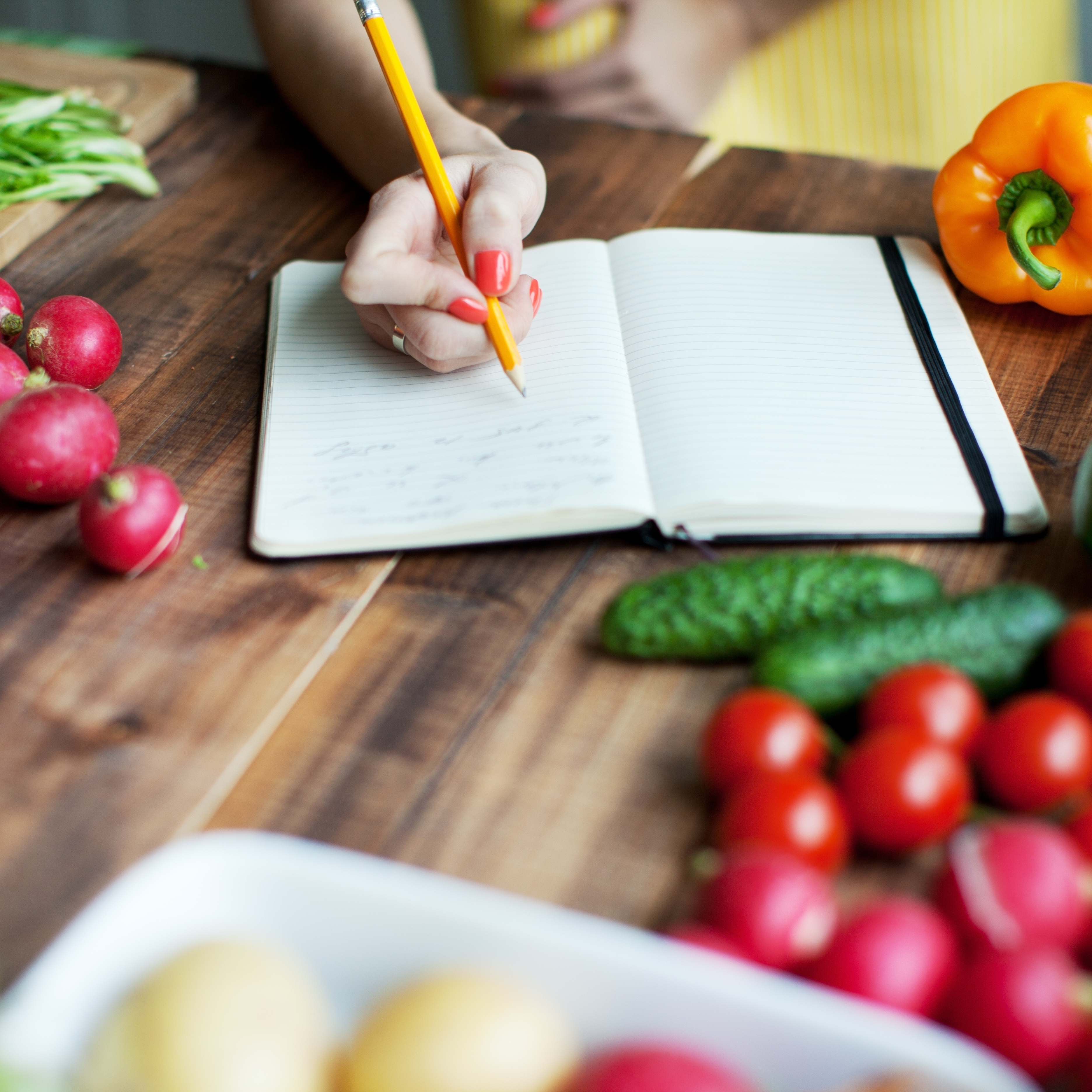 someone writing into a notebook, surrounded by fruit and vegetables