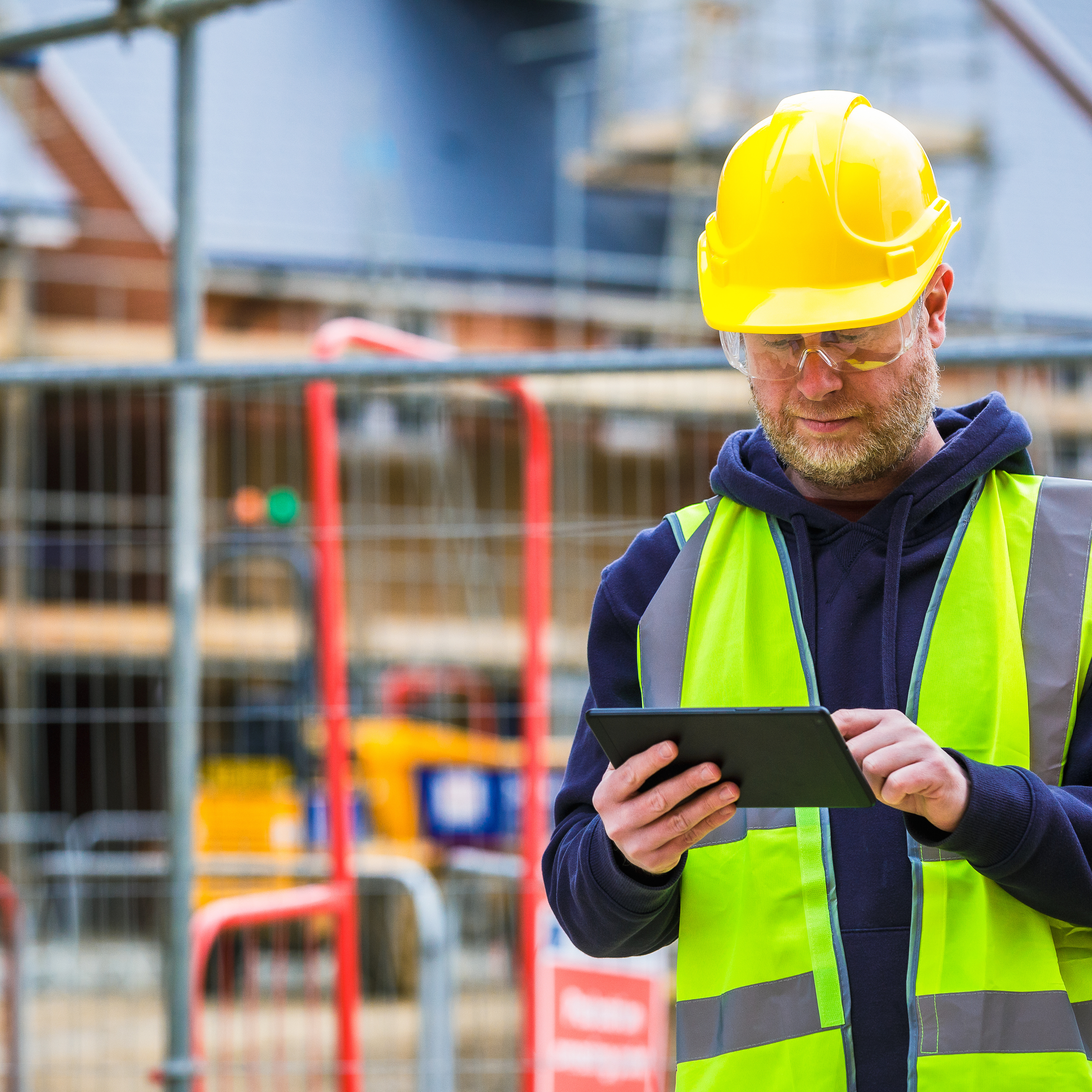 A man in hard hat and high-vis vest on a construction site, looking down at a tablet