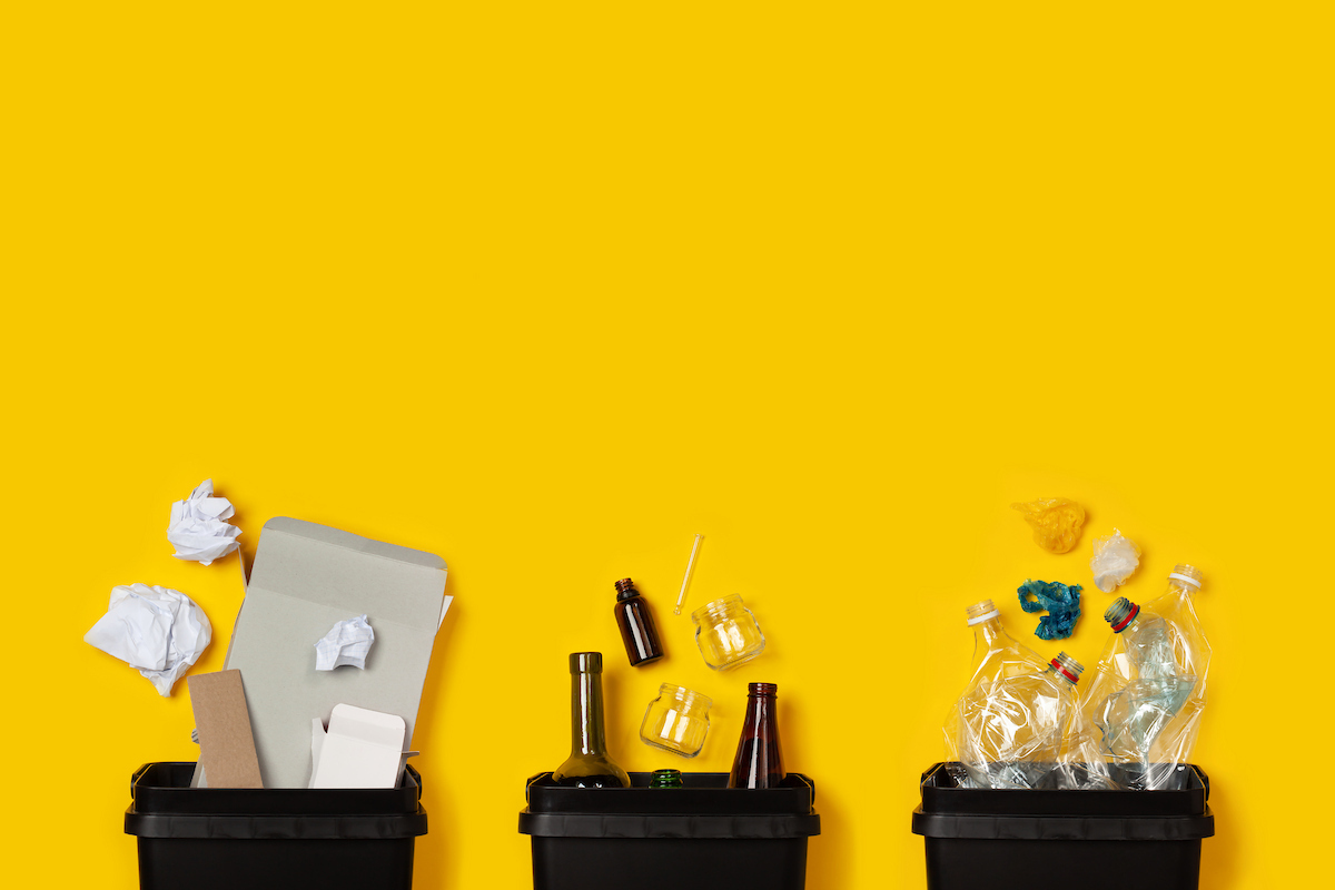Three recycling bins containing paper, glass, and plastic waste against a yellow background