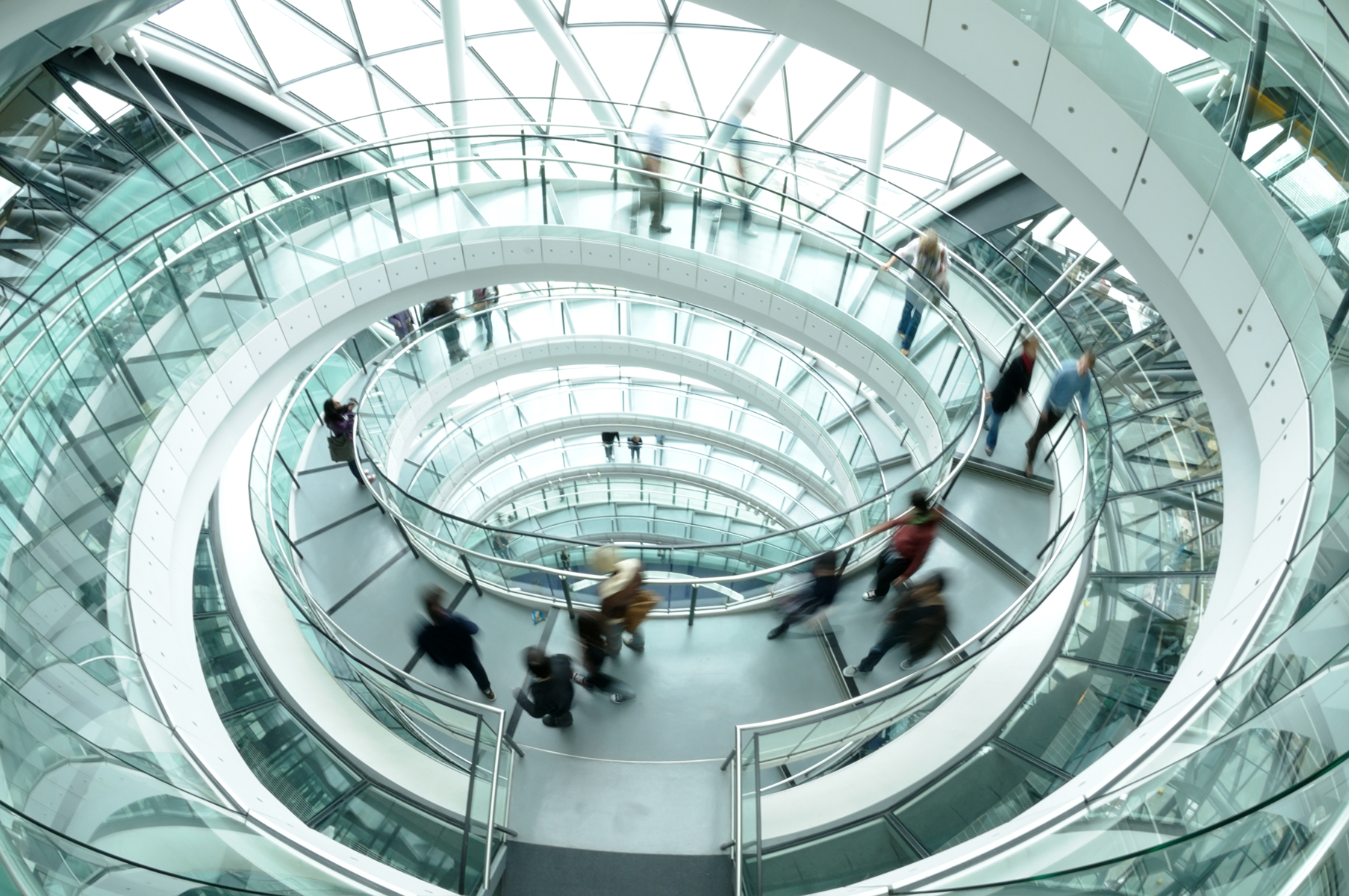 People walking along a circular, glass-walled spiral walkway inside a modern building.