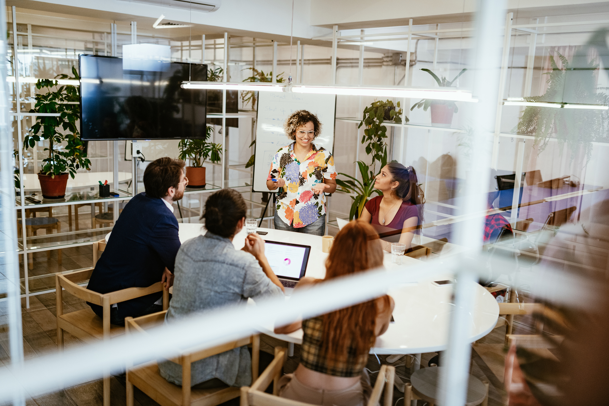 Person leading a discussion at a table while others listen and work in a bright meeting room.