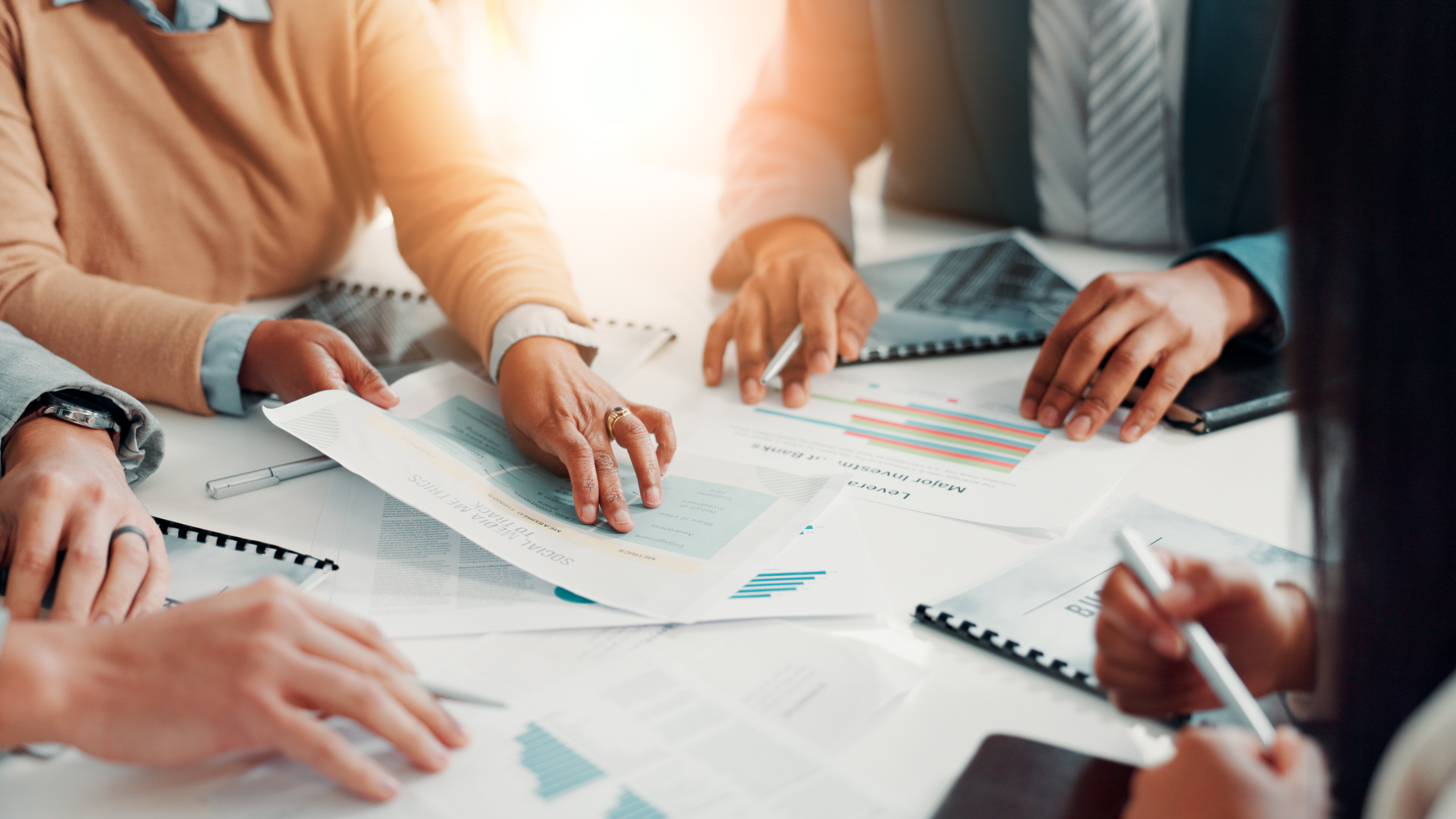 Hands of several people reviewing printed charts and documents around a meeting table.