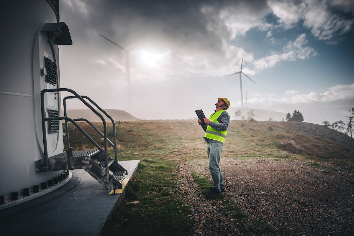 Technician in safety vest inspecting equipment at a wind farm with turbines in the background.