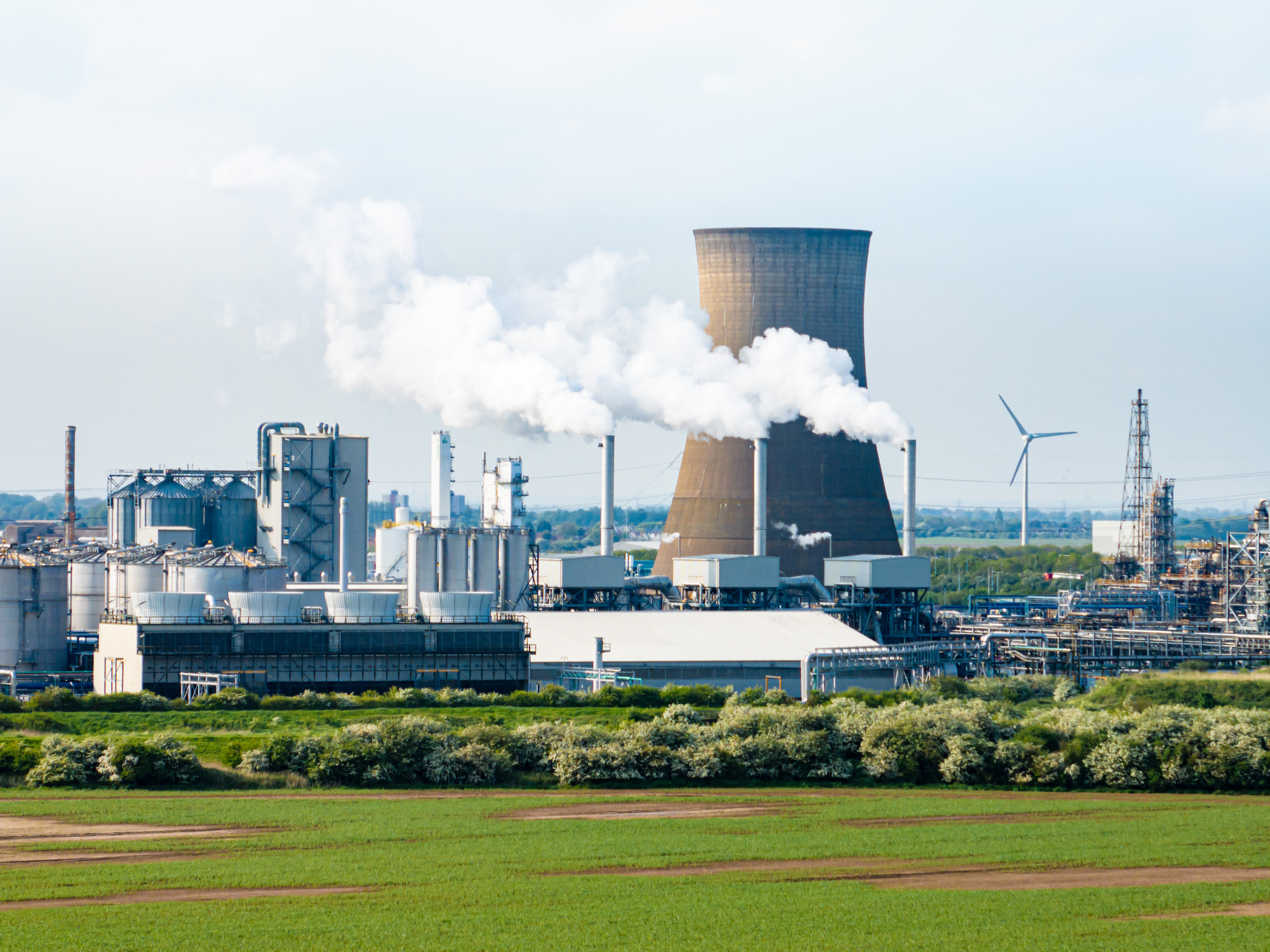 Industrial plant with cooling tower and chimneys emitting steam, with a wind turbine in the background.