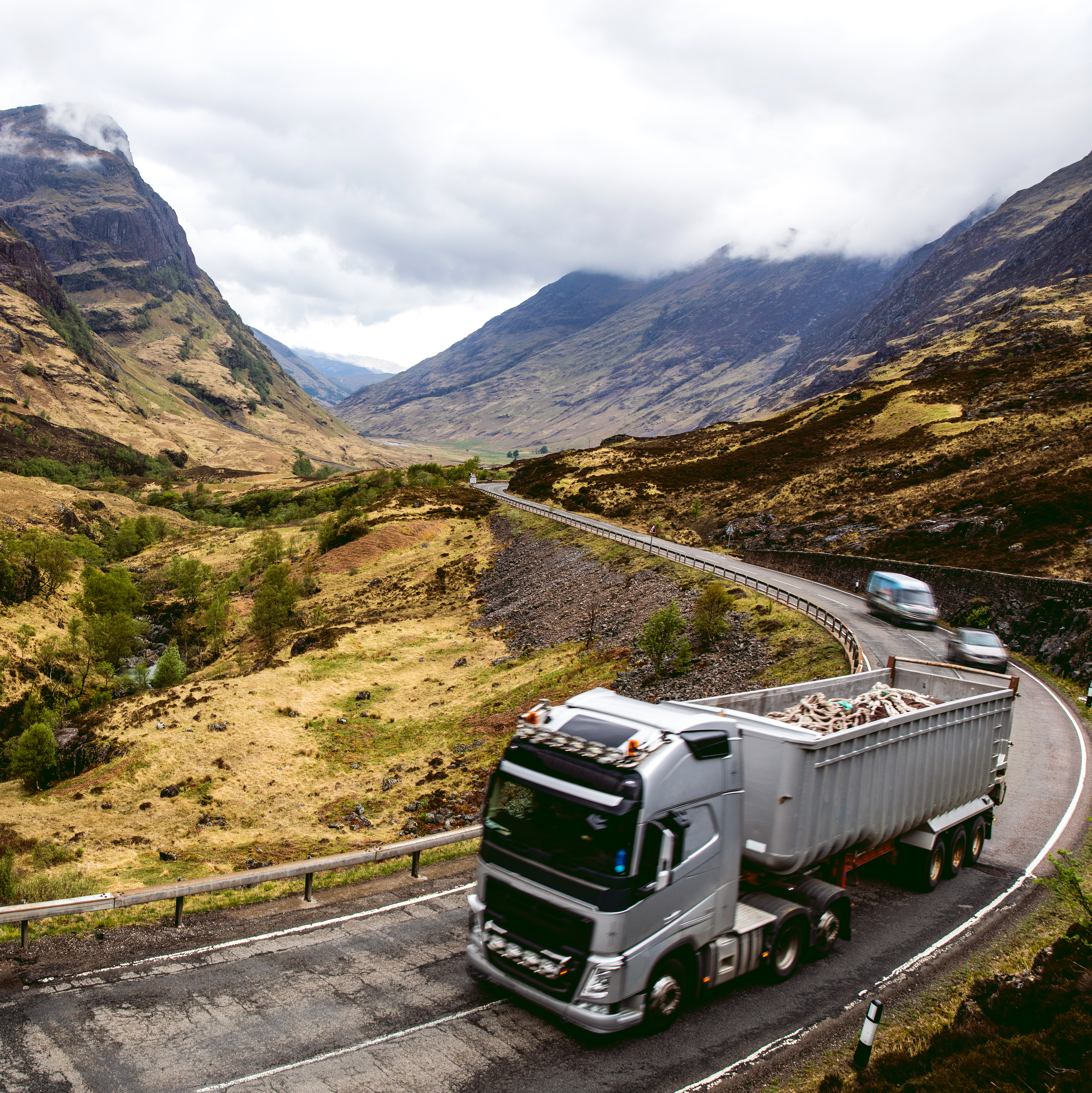 Mountain valley in Scotland with a winding road and a truck driving through rugged hills under low clouds.