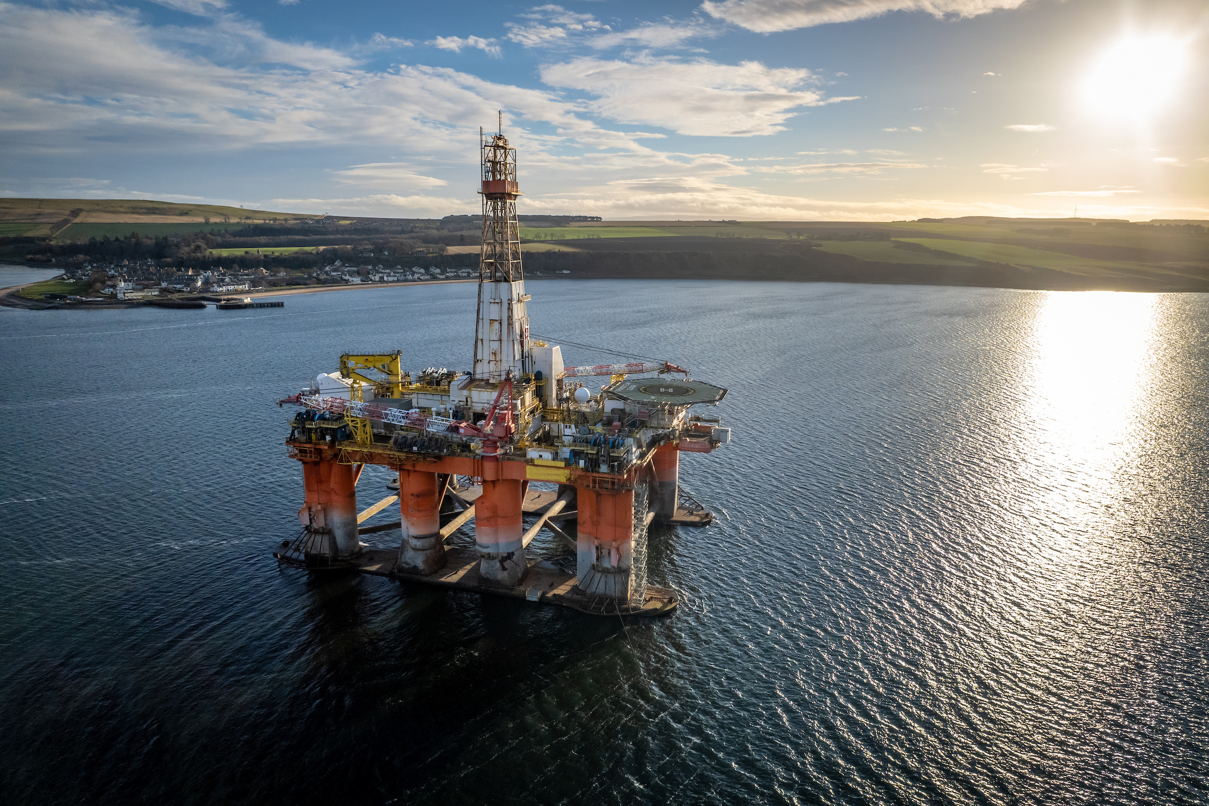 Offshore oil rig in calm water near a coastal village at sunset.