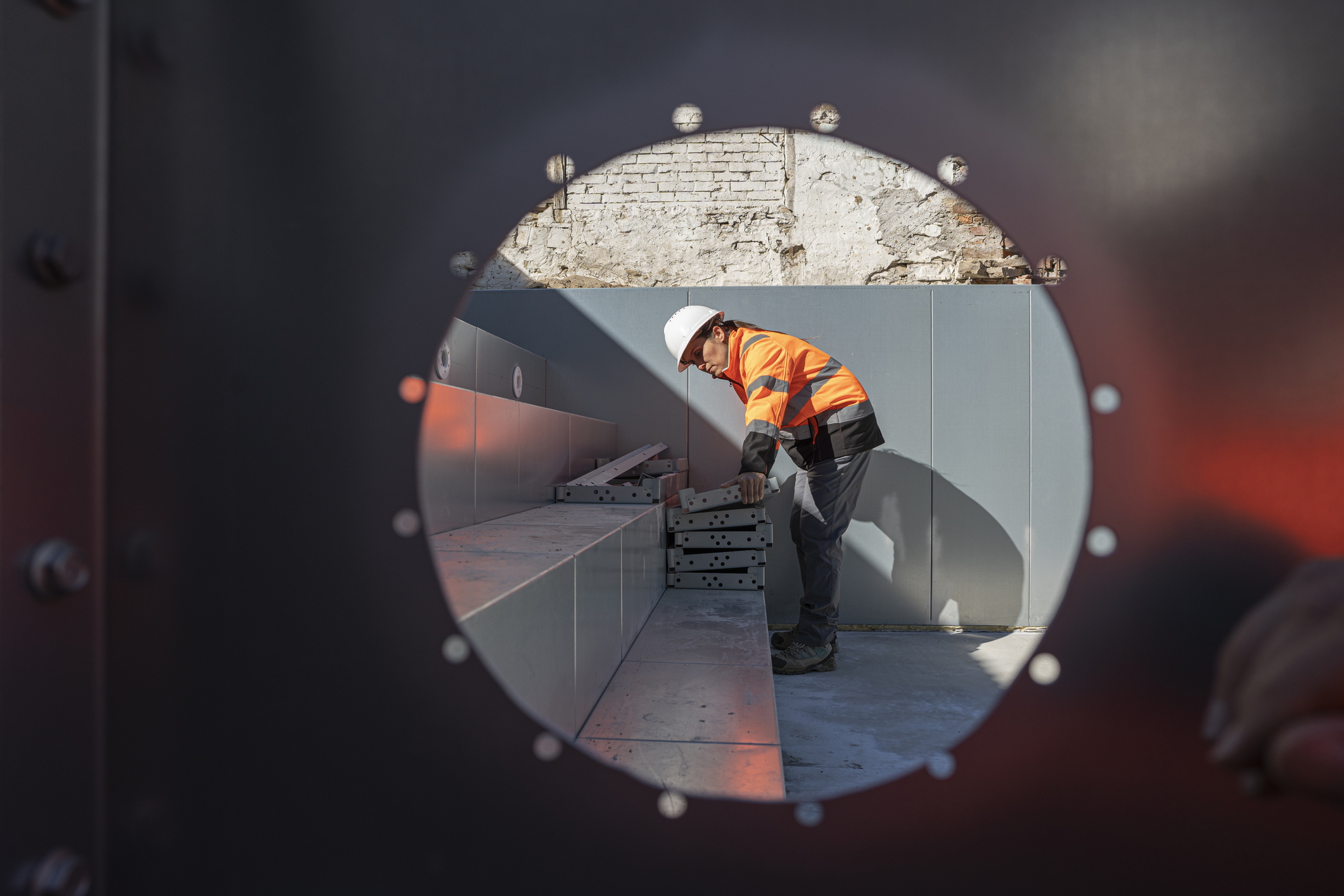 Construction worker in a high-visibility jacket and hard hat stacking metal components at a worksite, viewed through a circular opening in a steel structure.
